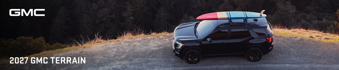 High-angle side view of a black GMC Terrain carrying a surfboard on a coastal cliff road.
