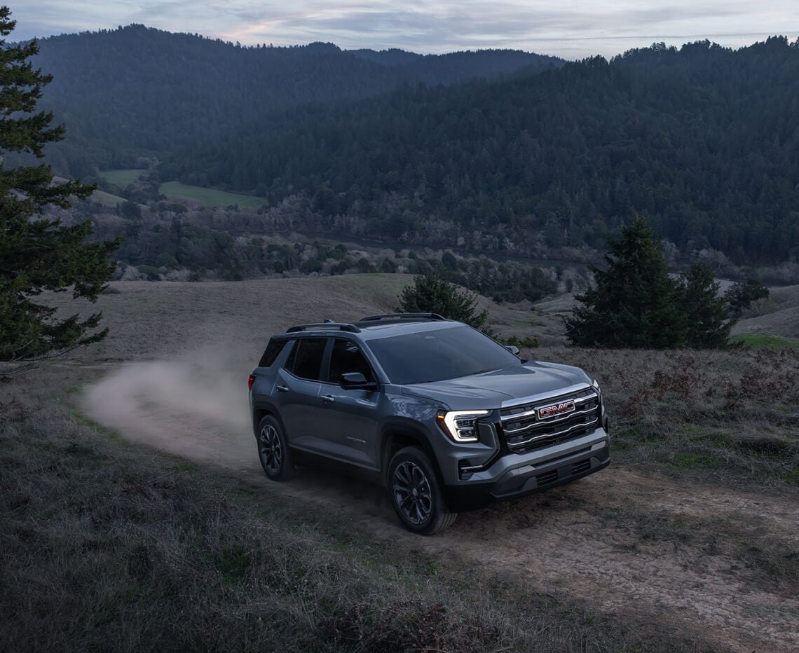 Front three-quarter view of a grey GMC Terrain driving on a dirt trail in grassy hills.