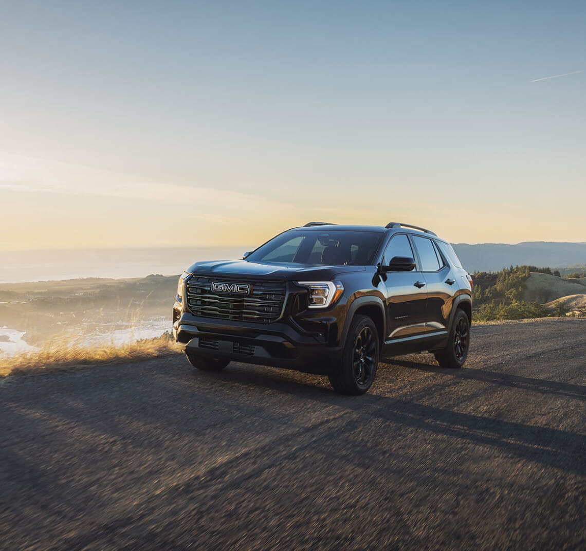 Front three-quarter view of a black GMC Terrain on a coastal mountain road at sunset.