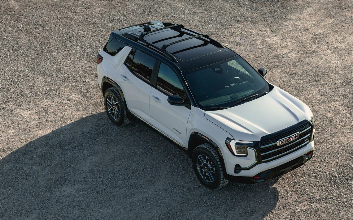 High-angle front three-quarter view of a white GMC Terrain AT4 with black roof on a gravel lot.