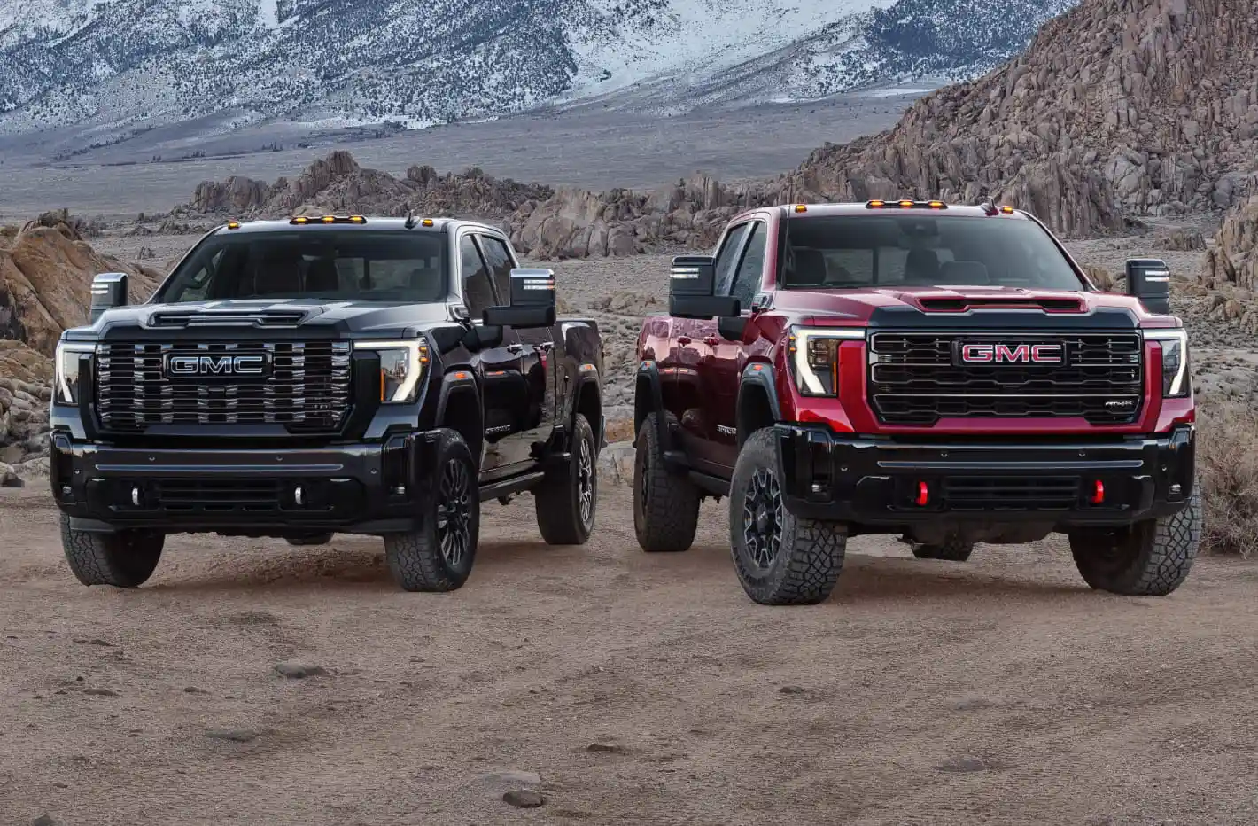 Featuring the 2026 GMC Sierra 2500 HD Denali Ultimate and 2026 GMC Sierra 2500 HD AT4X parked on a rocky desert terrain with snow-capped mountains in the background.
