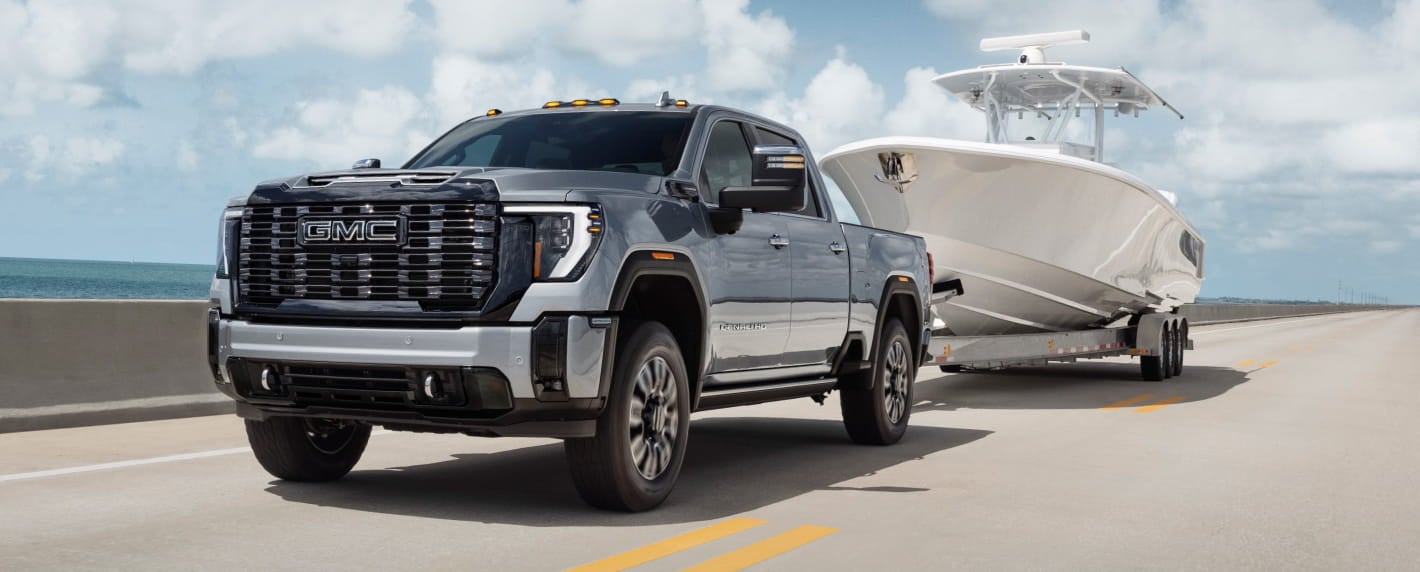 2026 GMC Sierra 2500 HD truck tows a sleek boat on a coastal highway under a blue sky with fluffy clouds.